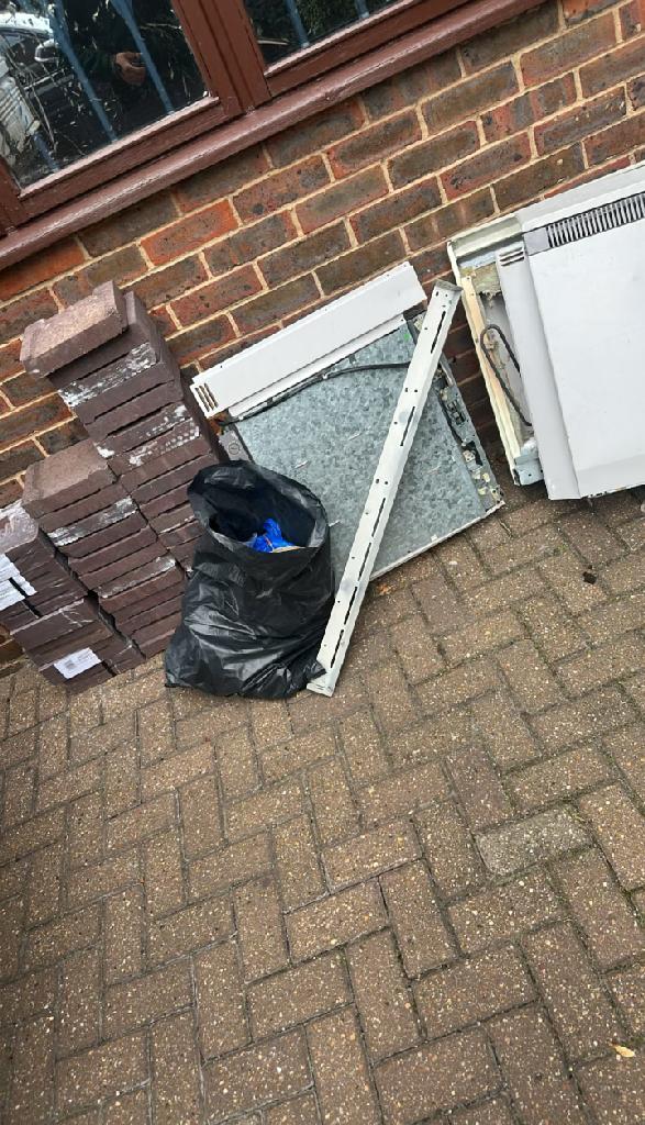 Before clearance in Bromley Town: a paved patio area along a brick boundary with a black bin bag and additional construction and debris items positioned near the wall.